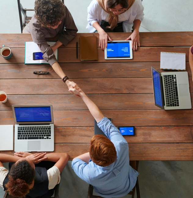 Gruppe von Geschäftsleuten an einem Holztisch bei einem gemeinsamen Meeting mit Laptops und Tablets, während zwei Teammitglieder sich die Hand geben, symbolisiert Solavitas Partnerschaft und Engagement für Vertriebskooperation